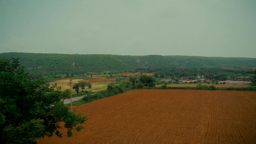 A serene, wide view of a plowed farm field in a lush green valley under a grey sky as a motorcycle drives by