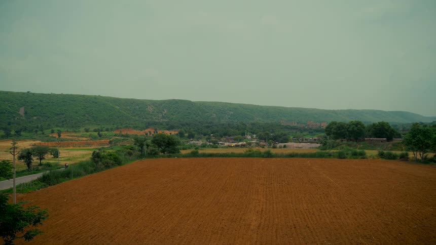 A wide shot shows a vast plowed farm field with lush green rolling hills in the background under an overcast sky