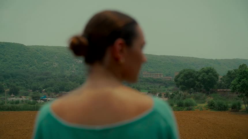 Over the shoulder rack focus shot of a woman with her hair in a bun looking out over a rural, hilly landscape