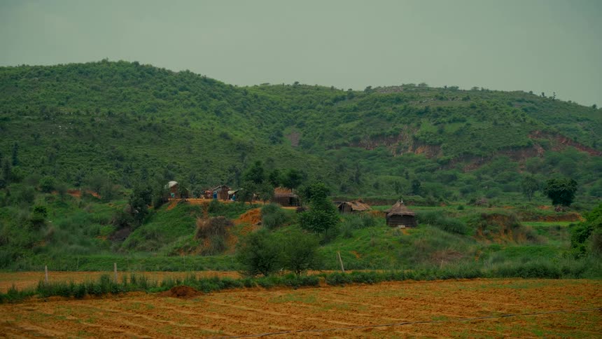A wide, static shot of a small rural village nestled among lush green hills with a tilled field in the foreground