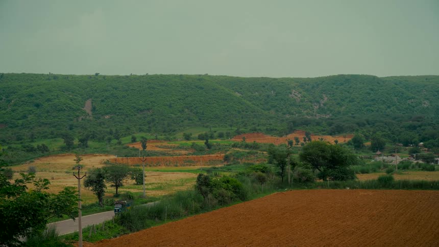 Vehicles travel along a rural road through a valley with plowed fields and lush green hills under a hazy sky