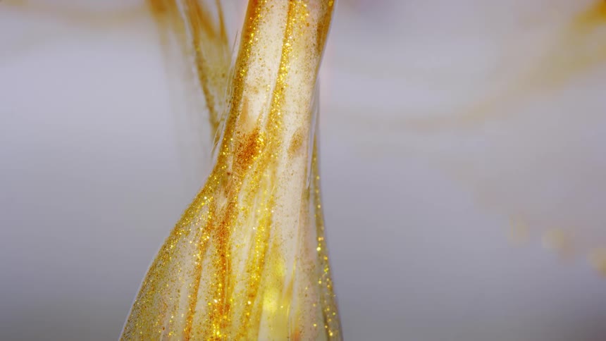 Golden glitter particles swirl hypnotically in a transparent, viscous liquid against a bright white background