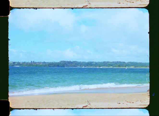 Vintage 8mm footage of a tranquil beach with gentle waves lapping the sandy shore and a distant, tree-lined coast
