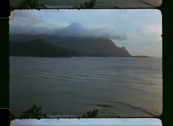 A vintage film time-lapse captures the sun setting over a calm ocean bay and a mountainous coastline