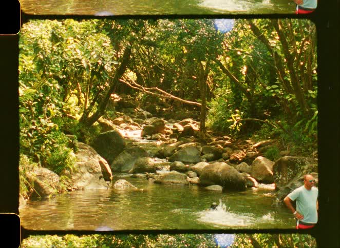Vintage 8mm film shows a person swimming in a secluded jungle river while a man watches from the rocky bank