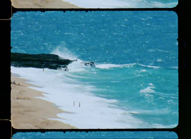 Vintage 8mm footage shows a wide shot of a powerful turquoise ocean with waves crashing against a sandy beach