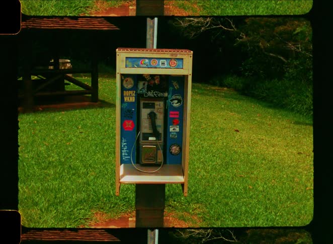 A vintage payphone covered in stickers and graffiti sits in a grassy park in this grainy film-style shot