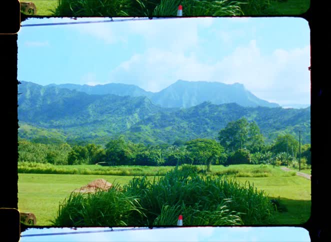 A vintage film shot of a serene, lush green valley with towering tropical mountains in the background