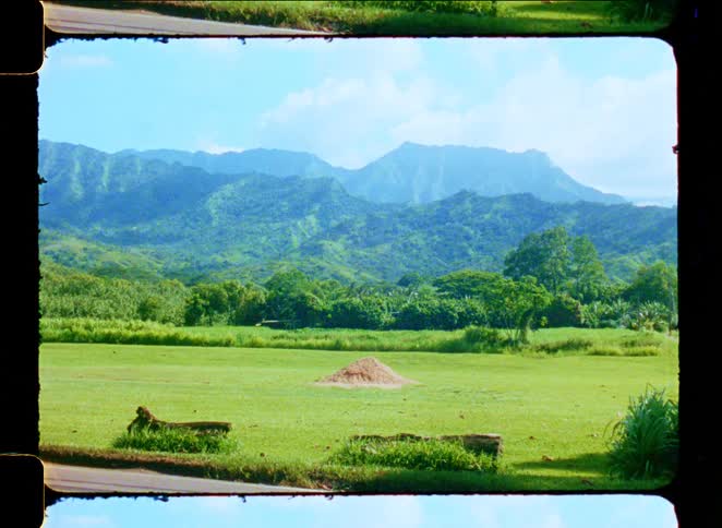 A vintage film shot captures a serene view of lush green mountains rising above a wide, open field