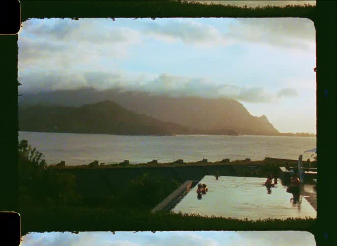 A vintage film shot shows people relaxing in an infinity pool overlooking a stunning mountain range by the ocean at sunset