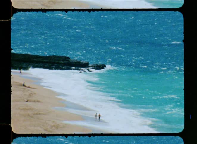 A vintage 8mm film captures a distant view of people walking along the shoreline as powerful turquoise waves crash onto a sandy beach