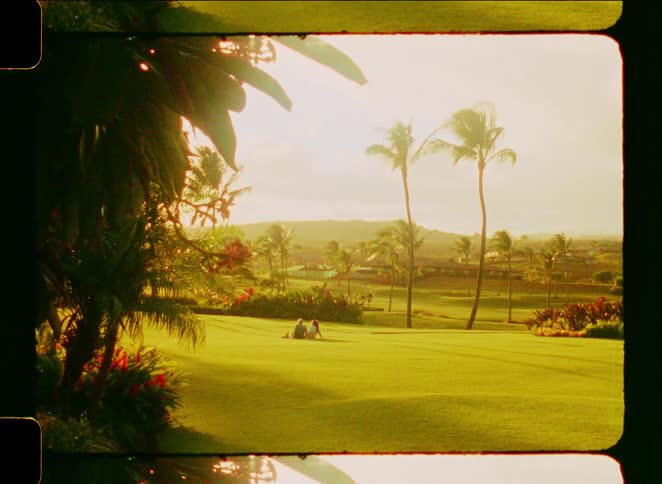 A nostalgic film-style shot of a couple relaxing on a vast green lawn in a tropical paradise with palm trees at sunset