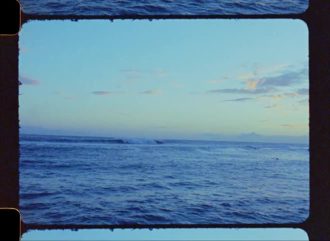 Vintage film footage shows surfers in the ocean waiting for waves to break during sunset