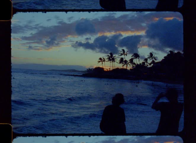 A vintage film frame captures the silhouette of a couple watching a tropical sunset over the ocean with palm trees