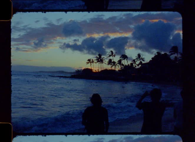 A vintage 8mm film-style shot captures the silhouette of two people watching a beautiful tropical sunset over the ocean waves