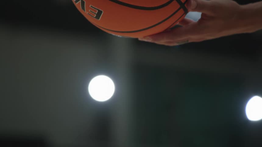 A dramatic out-of-focus close-up shows a basketball player dribbling in slow motion under bright gym lights