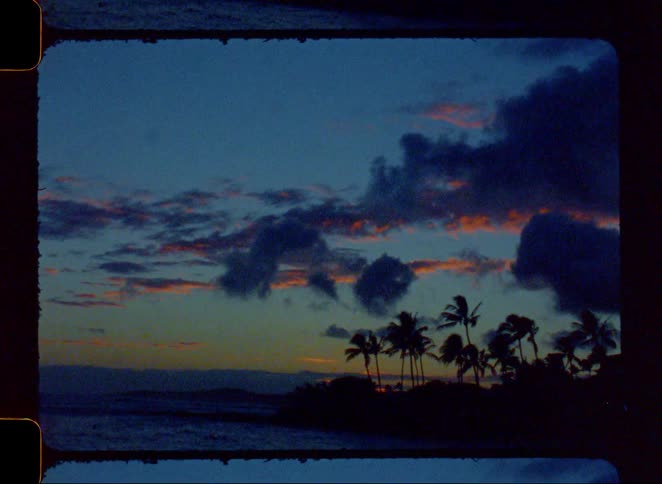 Vintage film captures a slow pan of a tropical sunset over the ocean with silhouetted palm trees