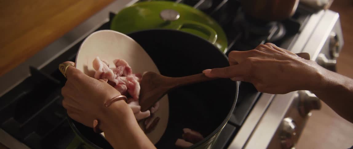 In slow motion, a chef carefully adds chopped raw pork to a large pot on the stove to start cooking a meal