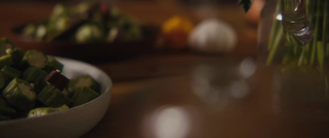 A slow pan across a rustic wooden table reveals a bowl of freshly chopped okra, with bread and herbs blurred in the background