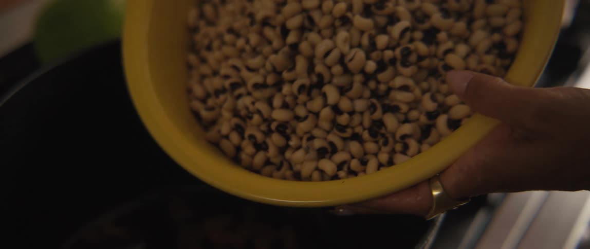 Slow motion close-up of a person pouring a yellow bowl of black-eyed peas into a pot on the stove to cook