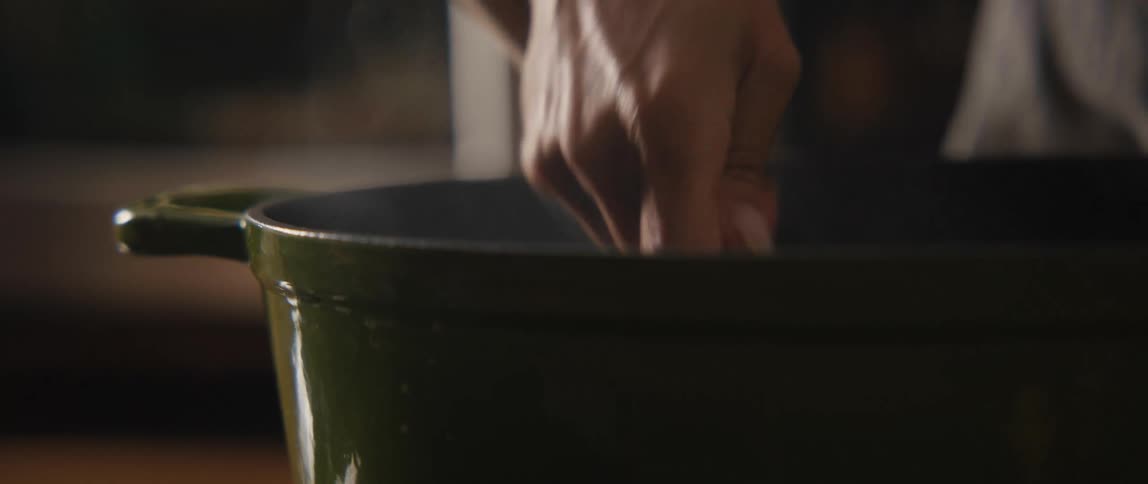 A hand stirs a steaming meal in a green pot with a wooden spoon in cinematic slow motion