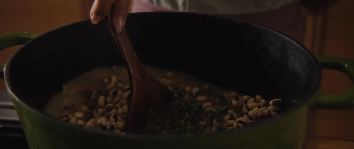 A person slowly stirs a simmering pot of black-eyed peas with herbs and spices using a wooden spoon