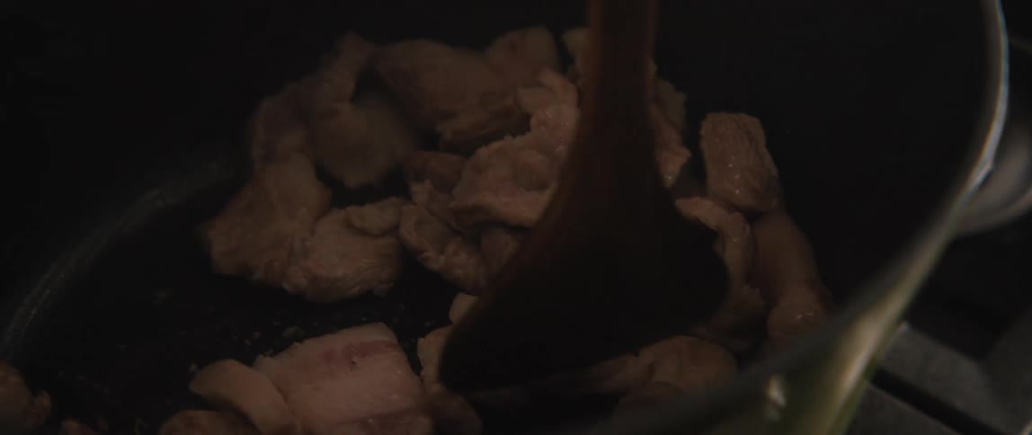 A cook stirs sizzling pieces of pork in a dark pot with a wooden spoon in slow motion