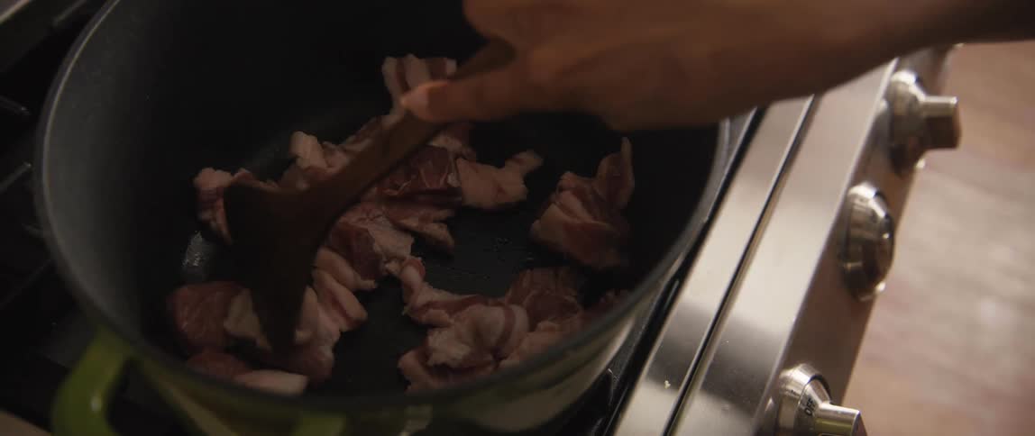 A person sautés diced pork belly with a wooden spoon in a pot on a gas stovetop