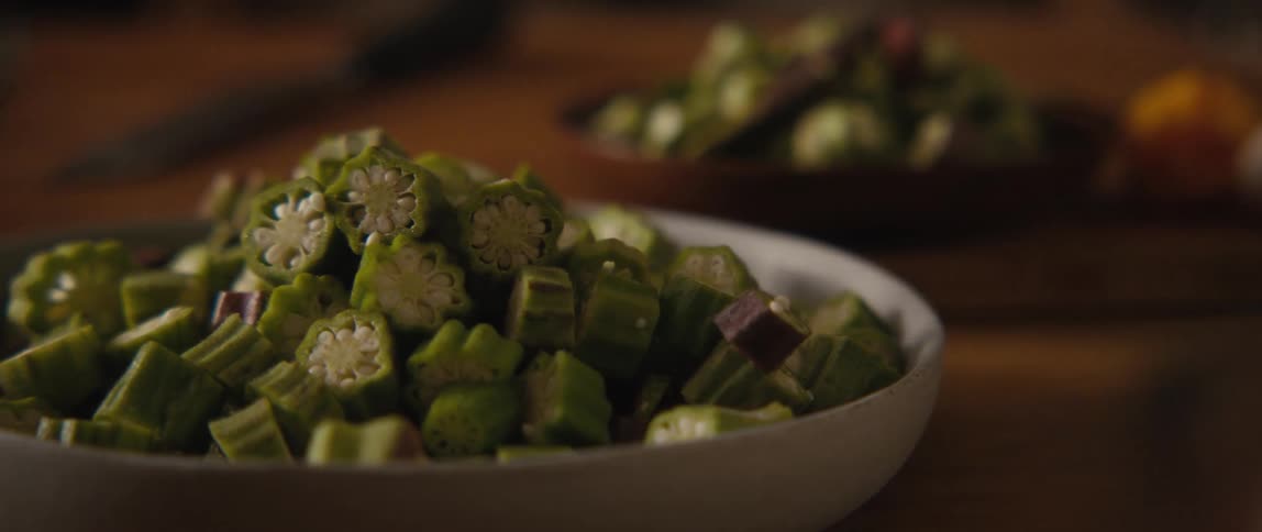 A cinematic slow-motion pan reveals a dinner spread with roasted pork knuckle, a vase of herbs, and bowls of freshly chopped okra on a wooden table