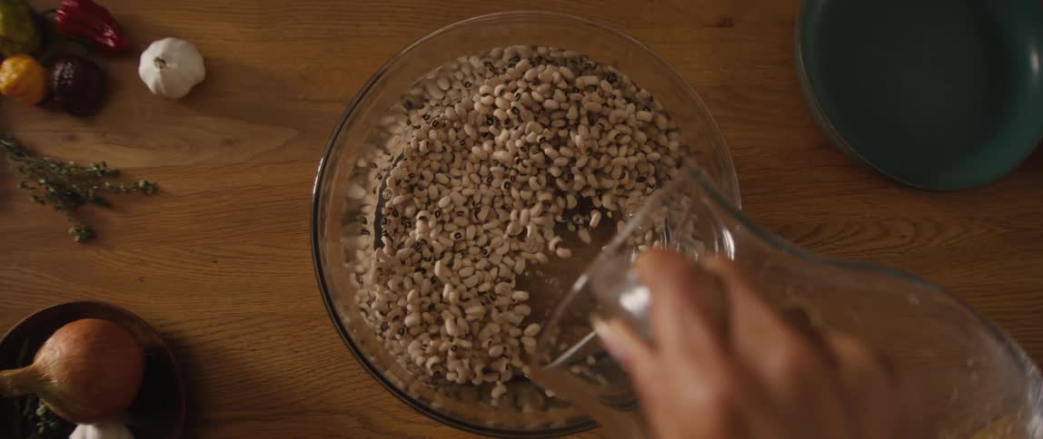 A top-down, slow-motion shot of water being poured from a pitcher into a glass bowl filled with black-eyed peas