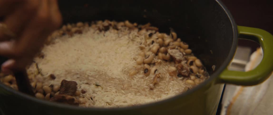A chef adds uncooked white rice to a simmering pot of black-eyed peas and meat, stirring the ingredients together with a wooden spoon