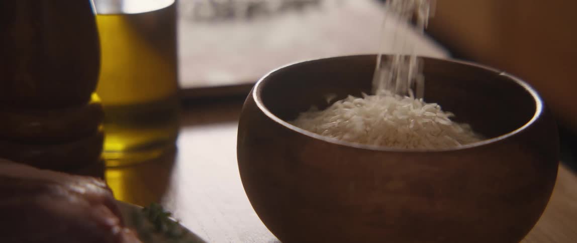 A steady stream of uncooked white rice falls into a rustic wooden bowl in slow motion