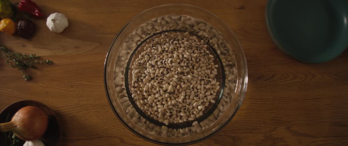 A top-down slow-motion shot of a hand gently washing black-eyed peas in a glass bowl of water on a wooden counter