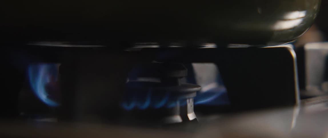 A gas stove burner ignites with a blue flame in a dark kitchen, shown in slow motion