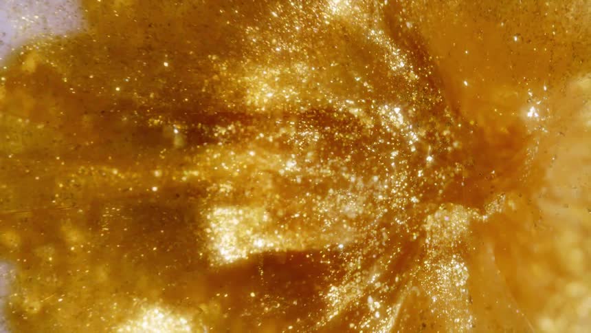 Shimmering gold particles dance and swirl within a thick, luxurious liquid in an extreme close-up shot