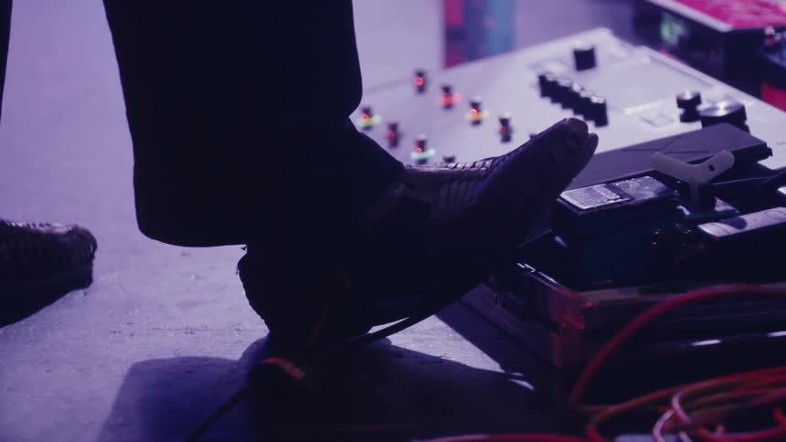 A musician's foot works a wah pedal on an effects board during a performance on a dimly lit stage