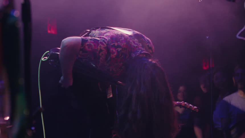 A guitarist with long hair headbangs in dramatic slow motion while performing on a purple-lit stage