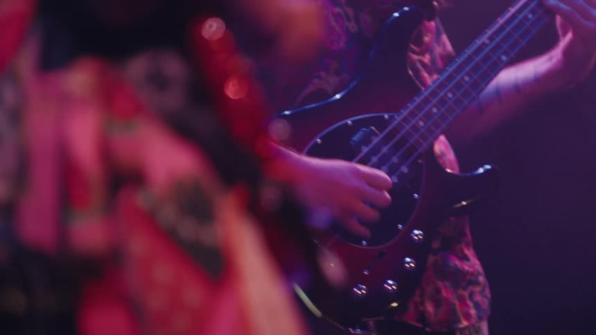 A rack focus shot reveals a long-haired bassist playing energetically on stage during a live show with purple and pink lighting
