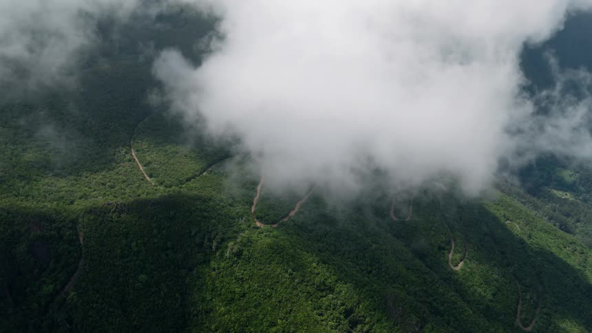 A static aerial view of a lush green mountain with a winding dirt road partially covered by thick clouds