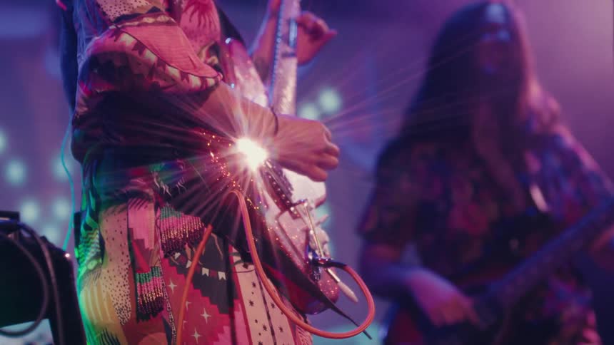 Close-up of a guitarist in a vibrant outfit playing a sparkly red electric guitar on a colorfully lit stage during a concert