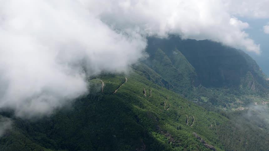 A tranquil and scenic view of a verdant mountain range with hairpin turns peeking through a layer of clouds