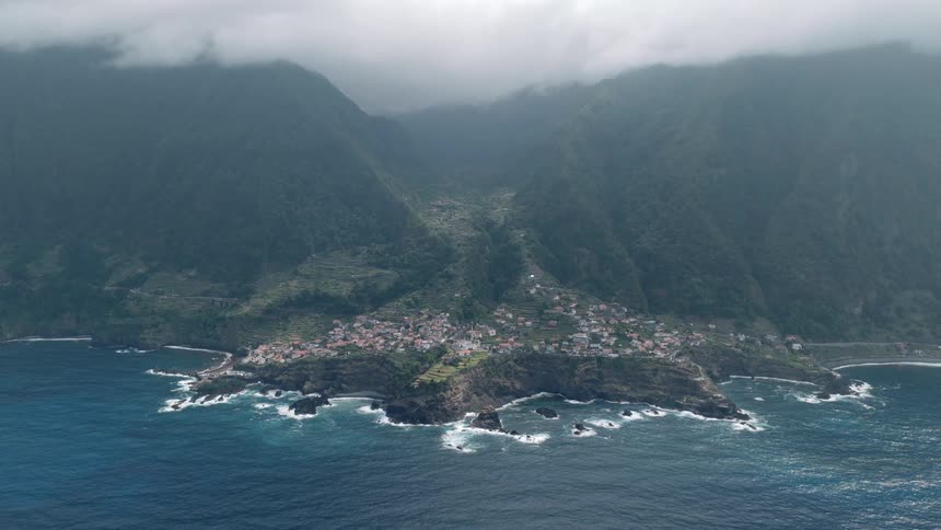 An aerial view of a remote coastal village nestled in a valley between lush green mountains and the rocky shore of the ocean on a cloudy day