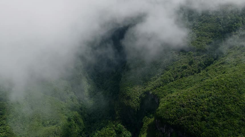 A static aerial view shows thick white clouds slowly clearing to unveil a deep, lush green mountain valley