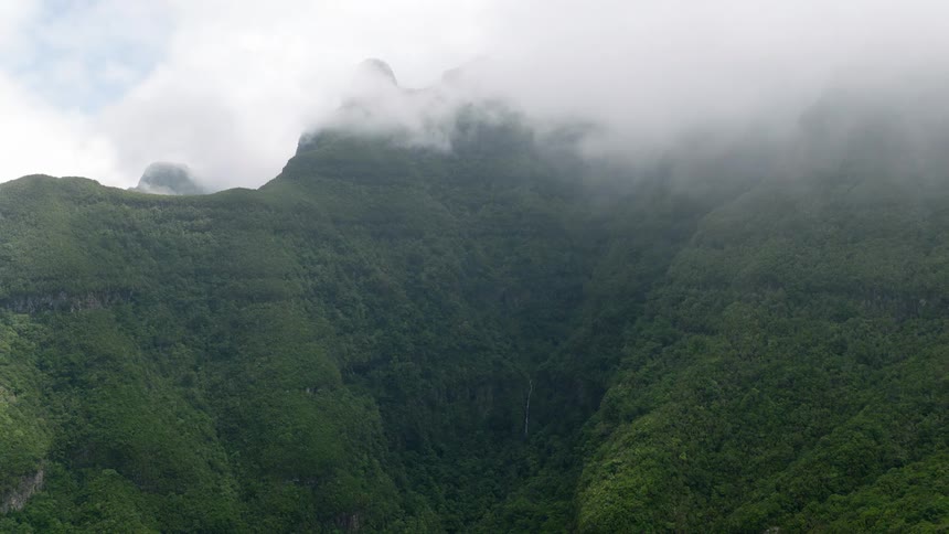 Aerial shot of a lush green mountain valley with a waterfall as clouds and mist slowly roll in