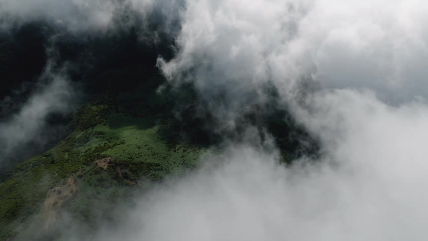 An aerial view from above shows a thick layer of white clouds slowly drifting over a lush green mountain landscape