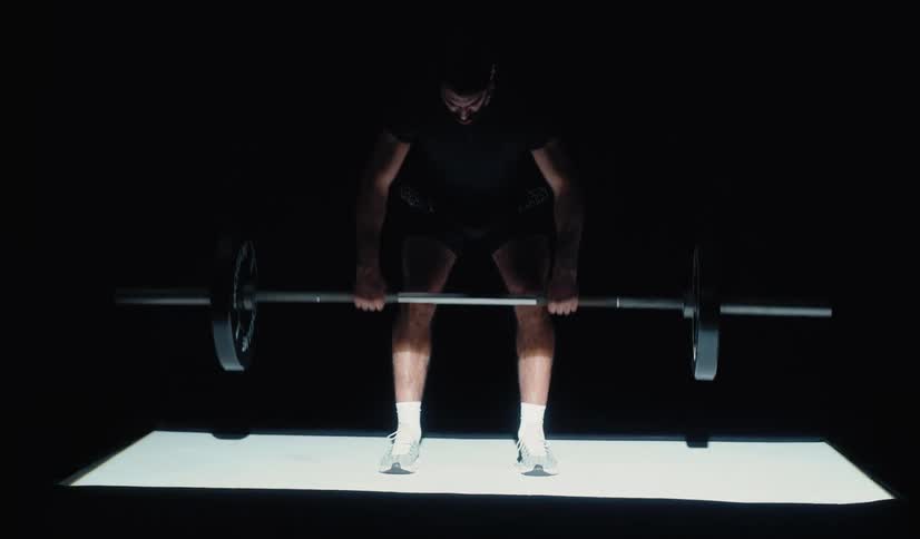 An anonymous man lifts a heavy barbell, doing bent-over rows in a dark spotlit studio