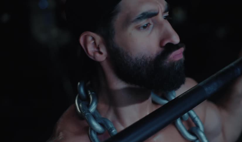 Intense close up of a muscular man with a beard and chain performing a pull-up in a dark gym