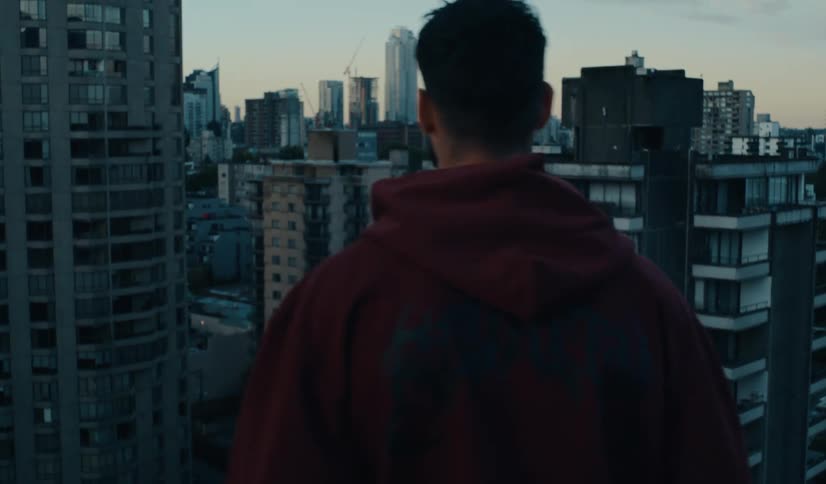 A man in a red hoodie stands on a rooftop looking out over the dense city skyline at dusk