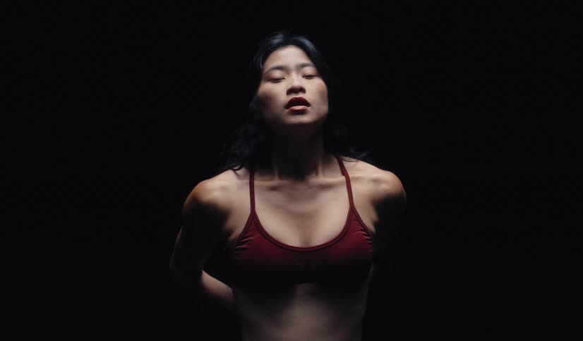 Slow motion of a strong Asian woman in a sports bra, she tilts her head back, then lowers it to stare intensely at the camera on a dark background