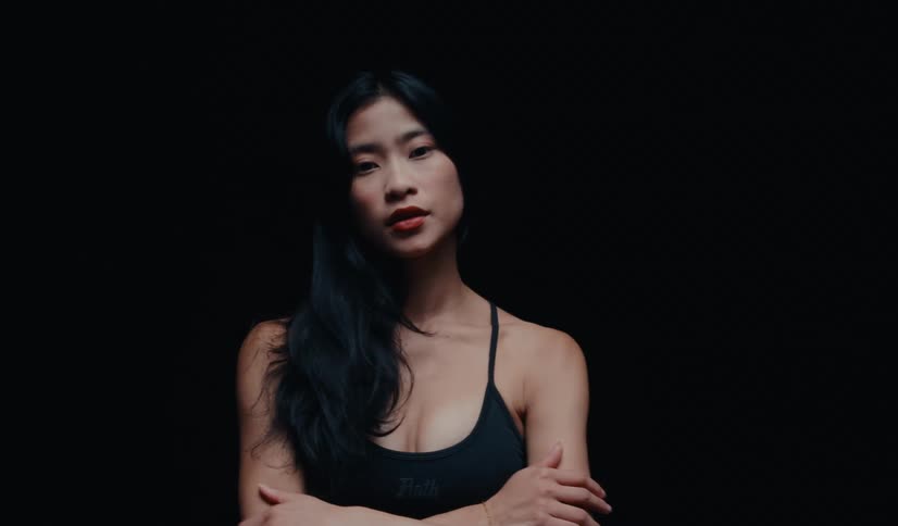 A confident and serious Asian woman in a black sports bra poses against a black background, slowly crossing her arms